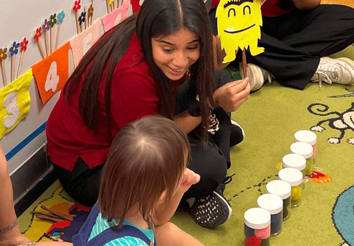 Smiling teacher holding yellow paper monster and small child looking with interest on her